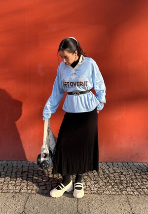 Mujer joven con una camiseta azul claro que dice "SUPÉRALO", falda negra, zapatos blancos, sosteniendo una bolsa, de pie contra una pared naranja.