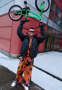 Kerékpár a fej felett tartva, élénk zöld vázzal, fekete gumik. A személy fekete pufi dzsekit, narancs-fekete mintás nadrágot és fehér tornacipőt visel.