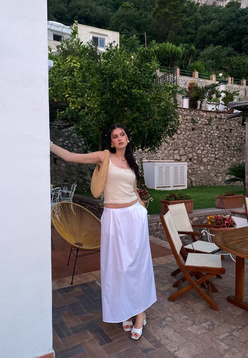 Ανοιχτό καφέ τοπ με υφασμάτινη υφή, λευκή φούστα μέχρι τον αστράγαλο, πλεκτή τσάντα και λευκά σανδάλια με χοντρό τακούνι σε εξωτερικό χώρο.