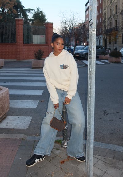 Sudadera crema de gran tamaño con gráfico de "Love Thrills", jeans anchos de tiro bajo en azul claro con efecto desgastado, y zapatillas negras, sosteniendo un bolso marrón.