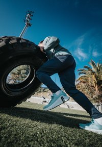 Une personne portant un sweat-shirt bleu sarcelle et un pantalon foncé pousse un grand pneu noir. Les alentours sont couverts d'herbe verte et de palmiers.