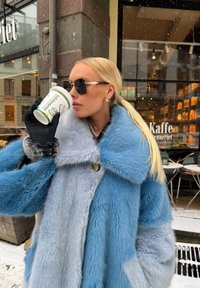 Απαλή γαλάζια και γκρι ψεύτικη γούνα μπουφάν με μεγάλο γιακά, κουμπιά και μαύρα γάντια. Το άτομο κρατάει ένα λευκό φλιτζάνι με πράσινα γράμματα.