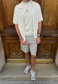 Camisa blanca texturizada de manga corta sobre una camiseta de color crema claro, combinada con pantalones cortos grises claros a rayas y zapatillas deportivas grises. Fondo de madera.