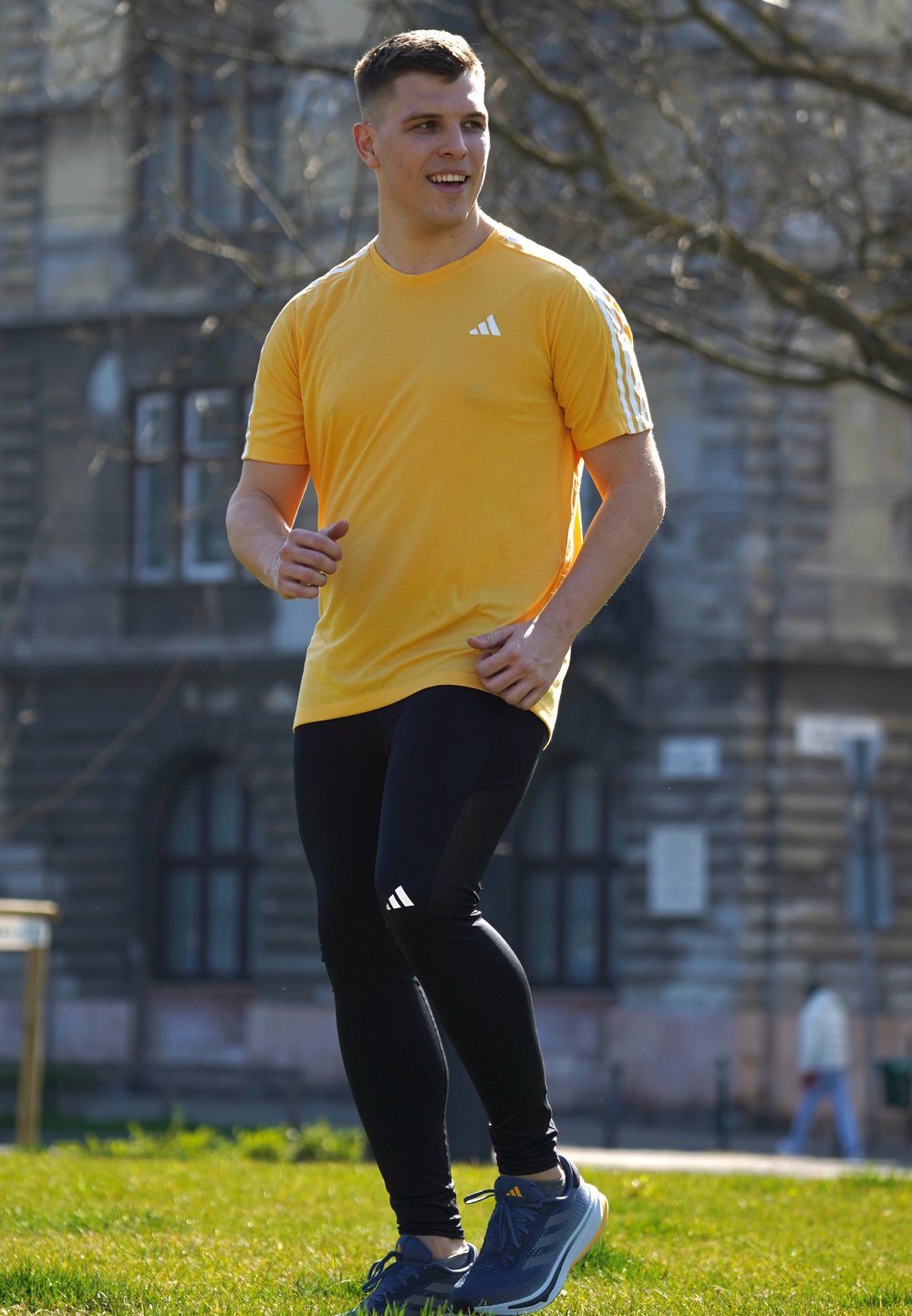 Rumena atletska majica s tremi belimi črtami na rokavih, v kombinaciji z črnimi leggins, ki imajo stranske akcente, in modrimi atletski čevlji.