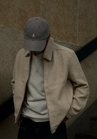 Beige gesteppte Zip-Jacke mit Kragen, getragen über einem hellen Hemd. Eine graue Mütze mit kleinem Logo. Neutraler Hintergrund.
