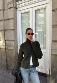 Mulher com um casaco com fecho verde escuro, jeans azul claro, óculos de sol pretos, segurando uma bolsa preta, parada contra uma parede de pedra bege e uma porta branca.