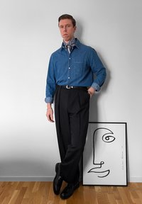 Chemise en denim bleu, pantalon plissé noir et chaussures noires. Une écharpe à motifs et une œuvre d'art abstraite en lignes sont placées à côté de la silhouette.