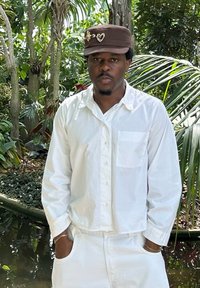 Witte overhemd met knoopsluiting en een borstzak, gecombineerd met witte broek. Bruine pet met decoratieve accenten. Weelderige groene achtergrond met loof.