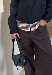 Veste marine portée sur une chemise à rayures blanches et brunes. Pantalon marron à jambes larges. Sac à bandoulière noir avec bandoulière et accents décoratifs.