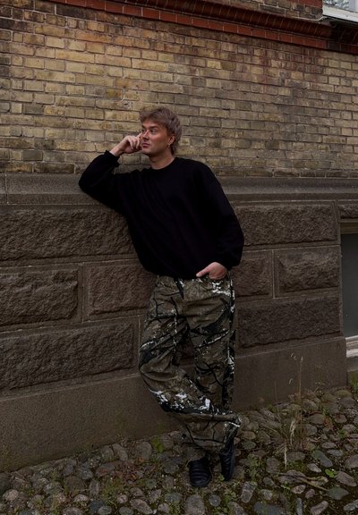 Suéter negro, pantalones con patrón de camuflaje y salpicaduras blancas, de pie frente a una pared de piedra texturizada, con calzado negro.