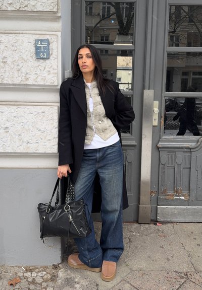 Mujer con cabello largo y oscuro que lleva un abrigo negro, chaleco beige, jeans anchos y zapatos color canela, sostiene un bolso negro en una calle de la ciudad junto a una puerta gris.