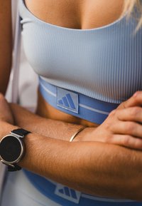 Sutiã desportivo listrado em azul claro com um logotipo na faixa, apresentando uma textura suave e um ajuste justo; relógio preto no pulso em primeiro plano.