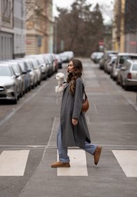 Femme en manteau gris traversant la rue, tenant un petit chien blanc, marchant sur un passage pour piétons en ville bordé de voitures garées des deux côtés.