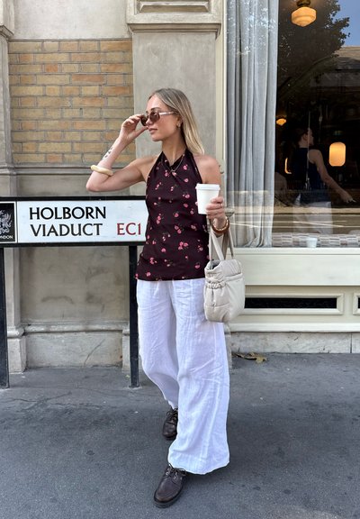 Vestida con un top halter de flores oscuras, pantalones de lino blancos y zapatos marrones con cordones, sosteniendo una taza de café y una bolsa color crema.