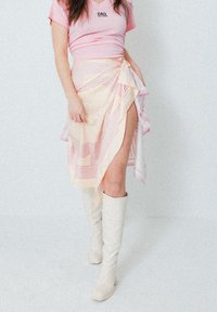 T-shirt rose à manches courtes avec logo, jupe enroulée rayée beige et rose à fente latérale, assortie avec des bottes blanches jusqu'au genou.