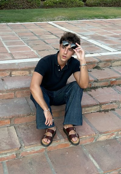 Camisa de polo negra, pantalones de mezclilla azul, sandalias marrones con hebillas, sentado en escalones de piedra, gafas de sol reposando en la frente, inclinándose ligeramente hacia adelante.