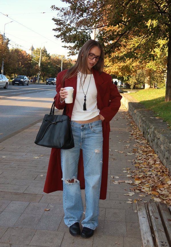 Une femme portant des lunettes, un manteau rouge et un jean déchiré tient un gobelet à emporter et un sac à main noir sur un trottoir urbain bordé d’arbres.