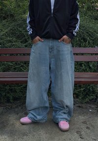 Φαρδιά ανοιχτόχρωμα μπλε τζιν με ξεθωριασμένη υφή, συνδυασμένα με παστέλ ροζ αθλητικά παπούτσια. Η φιγούρα φοράει ένα μαύρο μπουφάν με φερμουάρ.