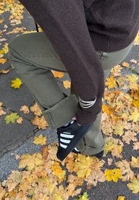 Mustat urheilutennarit valkoisilla raidoilla, yllään oliivinvihreät housut, istumassa maassa, joka on peittynyt keltaisiin syyslehtiin.