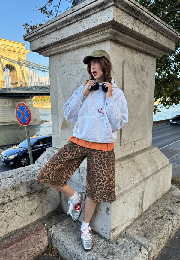 Persona joven con gorra, sudadera gris con capucha, pantalones cortos con estampado de leopardo y zapatos plateados apoyada en una estructura de piedra cerca de un puente y un río.
