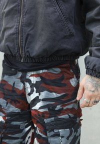 Must have musta bomberk jacket musta elastiku vööga, mis on kombineeritud punaste, hallide ja mustade kamuflaažiga cargo-lühkaritega, mis sisaldavad mitmeid taskuid ja omavad sileda tekstuuri.