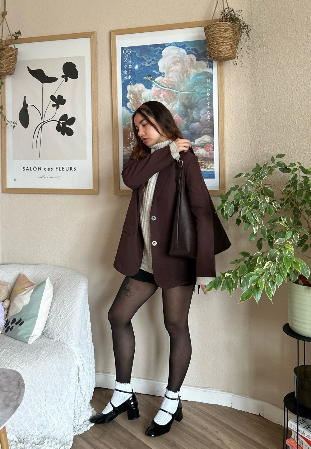 Brun blazer over en cremefarvet turtleneck, sorte shorts og mønstrede strømpebukser. Sort håndtaske, klodshæle og stueplanter i baggrunden.