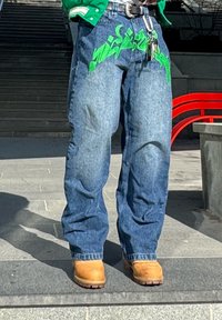 Άτομο που φοράει φαρδιά μπλε τζιν με πράσινο κείμενο σε στυλ γκράφιτι, καφέ εργασιακά μποτάκια, π ράσινο τοπ και κλειδιά κρεμασμένα από τη θηλιά της ζώνης σε εξωτερικό χώρο.