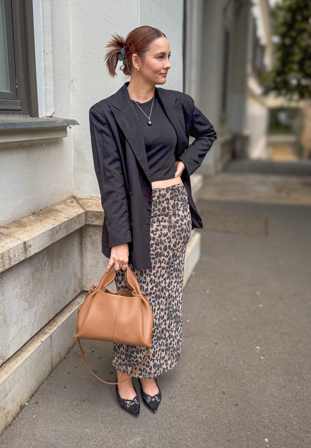 Μαύρο σακάκι, κοντή μαύρη μπλούζα, φούστα με λεοπάρ τυπωμένο σχέδιο, καφέ τσάντα, μυτερές μαύρες γόβες με λεπτομέρειες; εξωτερικός αστικός χώρος.