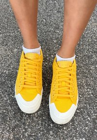 Κίτρινα καμβά sneakers με λευκές καουτσούκ σόλες, διαθέτουν τονικού χρώματος κορδόνια και λογότυπο στην γλώσσα και στα πλάγια. Φορεμένα με λευκές κάλτσες μέχρι τον αστράγαλο.