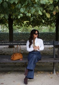 Chemise blanche à col, jean bleu délavé à jambes larges, sac à main en daim marron, écharpe marron et lunettes de soleil. Assise sur un banc dans un parc.