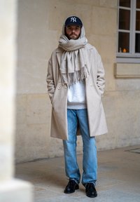 Cappotto di lana beige, maglione grigio, jeans blu e scarpe nere. L'individuo indossa un cappello scuro e una sciarpa beige chunky con frange.