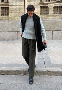 Pull gris clair à côtes, écharpe noire et pantalon vert foncé. Chaussures noires à texture lisse. Fond de trottoir urbain.
