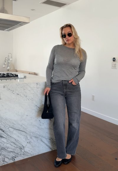 Mujer con gafas de sol, camiseta gris de manga larga y vaqueros oscuros apoyada en una encimera de cocina de mármol sosteniendo un bolso negro.