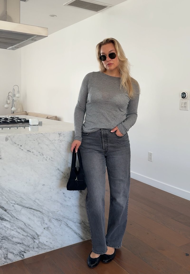 Γυναίκα με γυαλιά ηλίου, γκρι μπλούζα με μακριά μανίκια και σκούρο τζιν, στηρίζεται σε μαρμάρινο πάγκο κουζίνας κρατώντας μαύρη τσάντα.