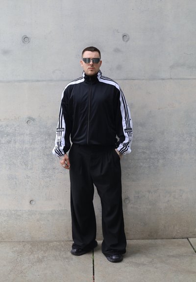 Chaqueta deportiva negra con rayas blancas, con cierre frontal, combinada con pantalones negros holgados. Unas gafas de sol reflectantes completan el conjunto.