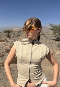 Ung kvinde iført beige ærmeløs top med lynlås og orange solbriller står i et stenrigt ørkenlandskab med sparsomt beplantede træer under en klar blå himmel.