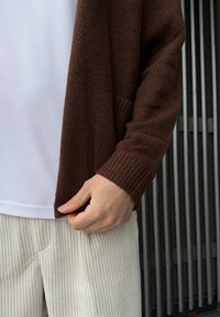 Casaco castanho de malha com punhos canelados e um bolso frontal, por cima de uma t-shirt branca e calças de bombazina bege claro. Detalhes do tecido texturizado visíveis.