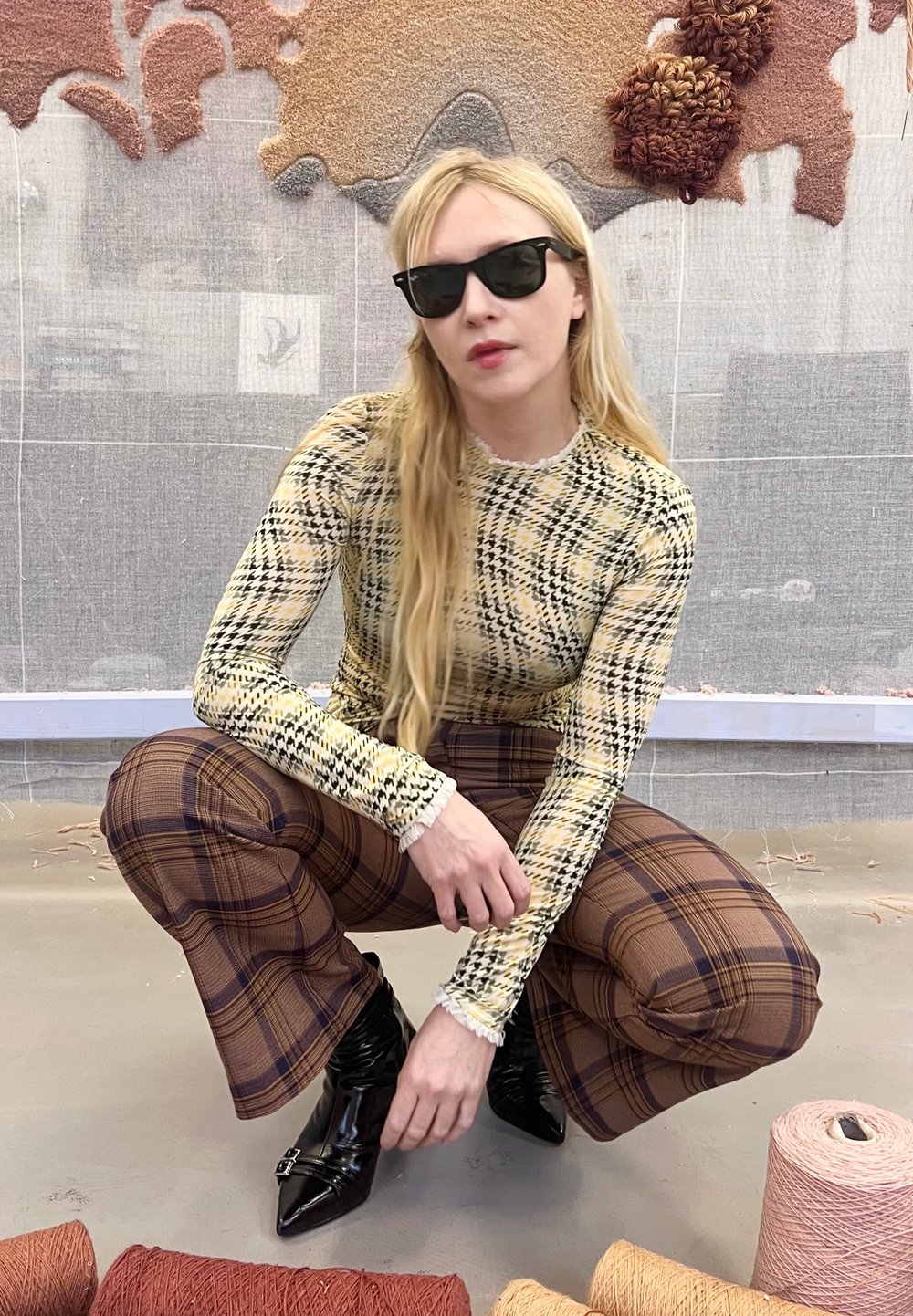 Donna con capelli biondi lunghi che indossa occhiali da sole, accovacciata, vestita con una maglia fantasia e pantaloni a quadri, circondata da rocchetti di filo e arte tessile.