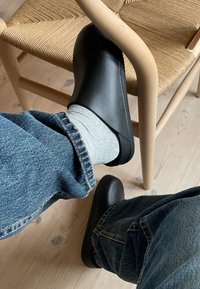 Zoccoli neri realizzati in materiale liscio con punta arrotondata, abbinati a calze grigie e jeans in denim blu. Una sedia in legno chiaro si trova sullo sfondo.
