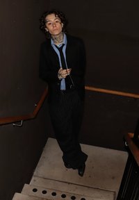 Persoană purtând un sacou negru peste o cămașă albastru deschis, pantaloni cu dungi, stând pe scări de beton cu balustrade din lemn și pereți întunecați.