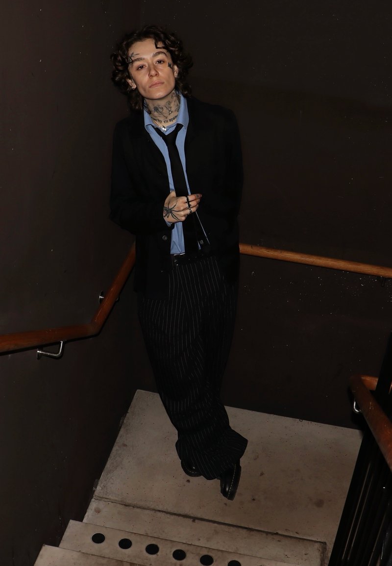 Persona che indossa un blazer nero sopra una camicia azzurra, pantaloni a righe, in piedi su una scala di cemento con ringhiere di legno e pareti scure.