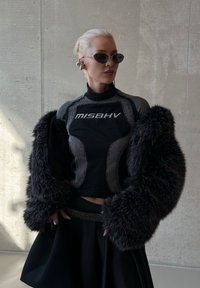 Femme blonde portant des lunettes de soleil noires, un haut MISBHV, de grandes manches en fausse fourrure noire, une jupe avec une ceinture cloutée, se tenant contre un mur en béton.