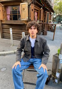 Άτομο καθισμένο σε καφέ καρέκλα, φορώντας σκούρο γκρι μπουφάν πάνω από μπλε πουλόβερ και ανοιχτό γαλάζιο τζιν, σε δρόμο με ξύλινες κατασκευές.