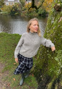 Grå, grovstrikket genser med høy krage, kombinert med et svart og hvitt rutete skjørt. Iført grønne gummistøvler. Omgitt av natur.