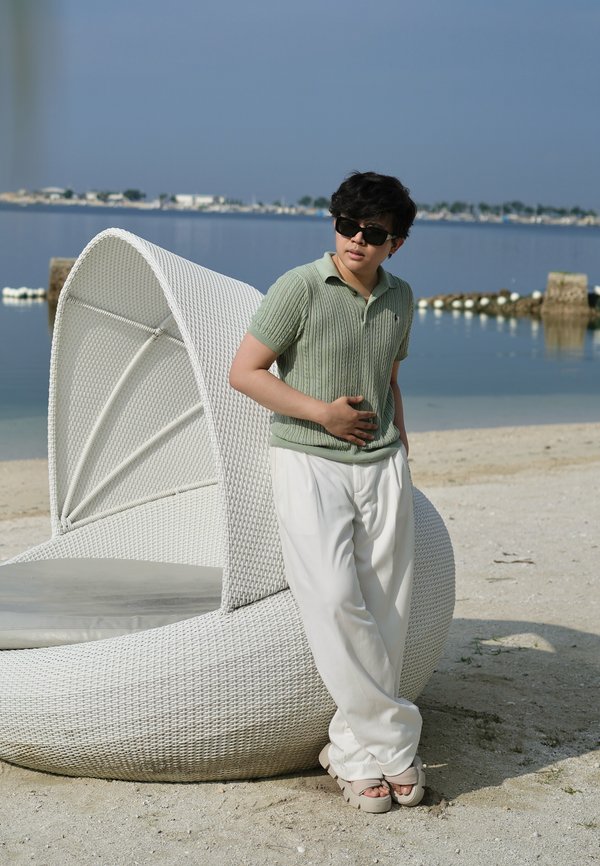 Giovane persona con occhiali da sole, maglione verde a maniche corte e pantaloni bianchi, appoggiata a una sedia a sdraio in vimini bianco vicino all'acqua calma della spiaggia.