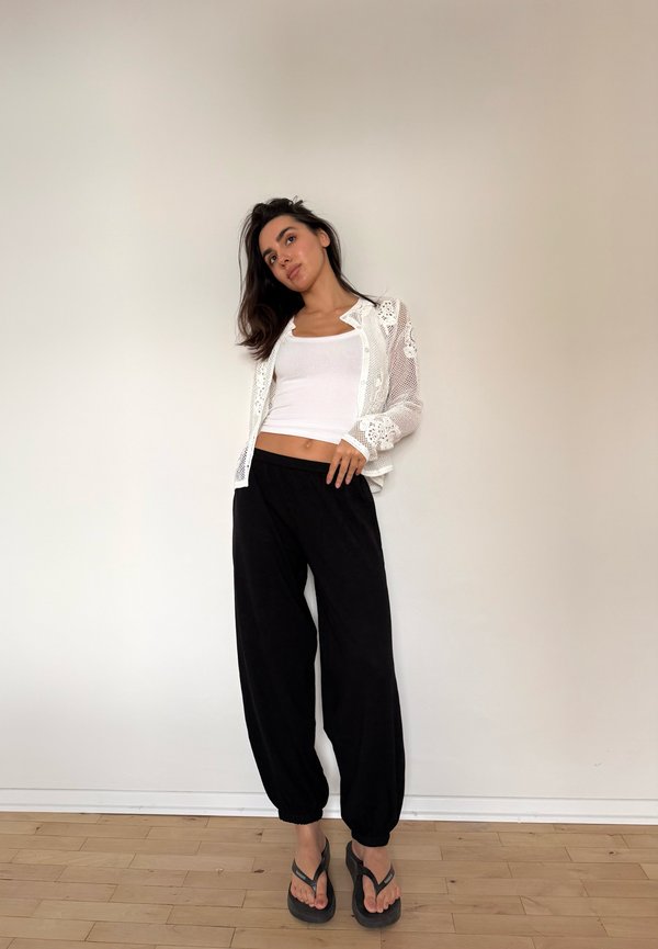 Woman with long dark hair wearing a white tank top, a sheer white lace shirt, black pants, and black flip-flops standing against a plain wall.