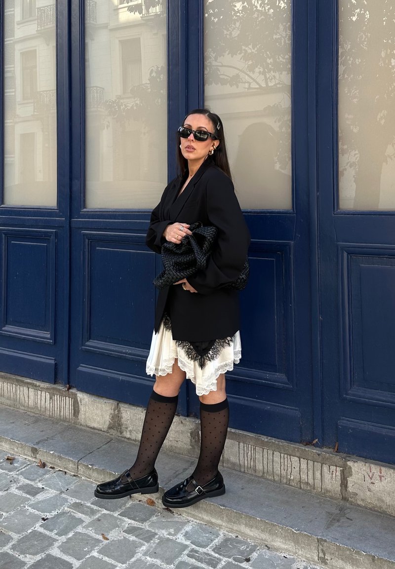 Blazer noir oversize sur une robe crème avec des bordures en dentelle noire, associé à des chaussettes hautes à pois noirs et des chaussures noires. Sac à main tissé inclus.