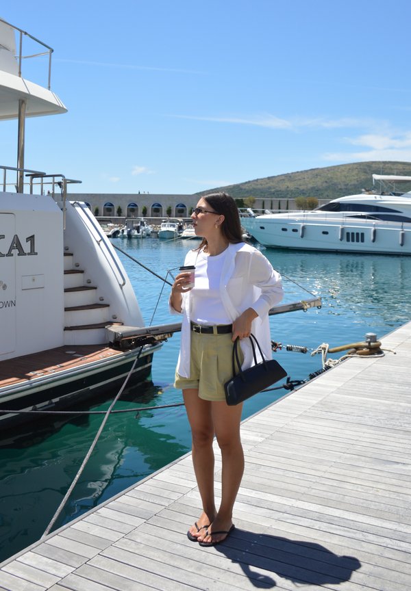 Femme portant des lunettes de soleil debout sur un quai de marina tenant une tasse de café et un sac noir près de yachts amarrés par une journée ensoleillée.