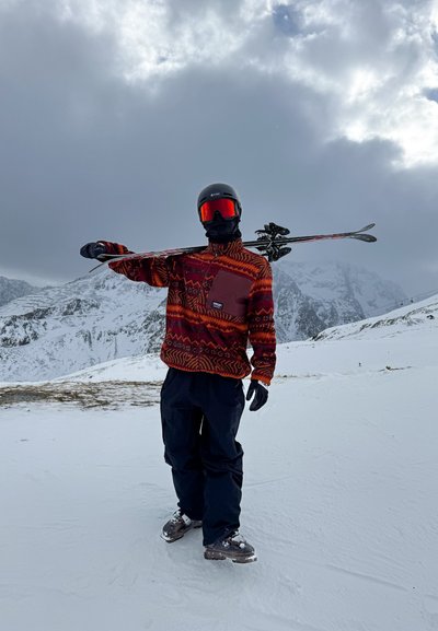 Chaqueta de forro polar con estampado rojo y naranja, pantalones de tirantes negros, guantes negros, casco de esquí y gafas, llevando esquís sobre el hombro en un terreno cubierto de nieve.