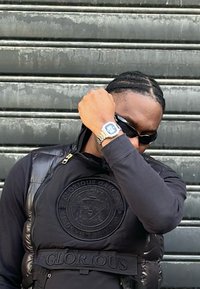 Sort puffervest med præget logo, båret over en skjorte med lange ærmer. Tilbehør inkluderer sølvur og mørke solbriller.